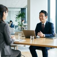 明るいオフィスの面接で、ジャケット姿の女性が、スーツ姿の面接官と向かい合って話している